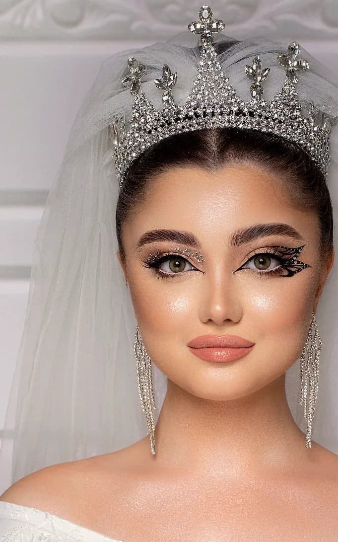 A tall crystal tiara over a veil, with a graphic liner and drop earrings